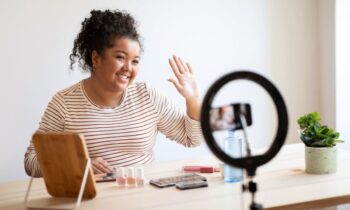 A content creator sitting a table, filming herself with makeup using a phone and ring light.