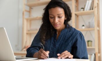 A woman working remotely from her home office, using a laptop and writing in a notebook.