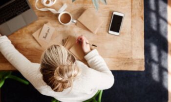 An overhead view of a woman working from her home office desk, using a laptop and writing in a notebook.