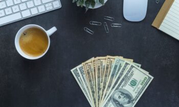 A remote worker's home office desk, with a computer, cash, coffee, and a plant sitting on it.