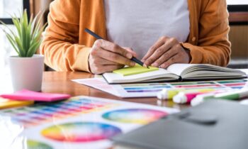 A freelance graphic designer working at her home office desk and writing in a notebook.