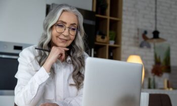An older woman in her home office, using a laptop and writing in a notebook looking for companies that hire older workers