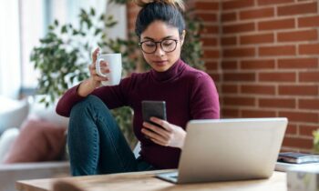 Woman sitting at her home office desk, using her phone while drinking coffee.