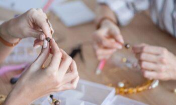 Closeup image of a woman's hands, holding beads and making jewelry at a large craft table.