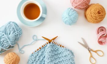 An overhead view of a set of knitting needles, an in-progress scarf, coffee, and a few extra skeins of yarn.