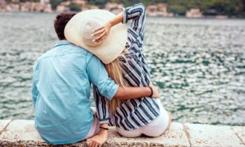 A couple sitting together on a rock wall, overlooking a lake with hills and buildings in the distance for blog post romantic business ideas