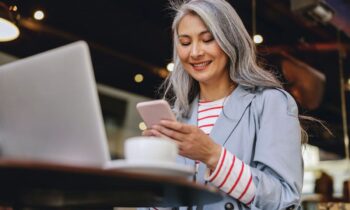 Woman sitting at a table in a cafe, using a smart phone and a laptop to work remotely for blog post work from home jobs that provide equipment
