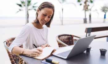 A woman working remotely on a laptop from a cafe table, with a beach and palm trees behind her.