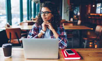A college-aged female with glasses in a cafe reading an article titled online jobs for college students on her laptop