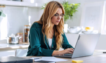 A woman with long blond hair and glasses reading an article on her laptop titled how to work from home as a bookkeeper