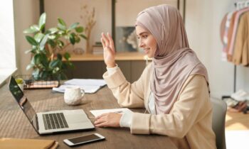 A woman using a laptop to take a video call from her home office, smiling and waving at the camera for blog post informational interviews to help land a job