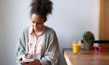 A woman sitting at home at a kitchen table, writing in a notebook thinking of ways to create an online writing portfolio.