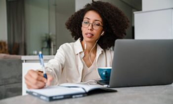 A woman working from home at her kitchen table, using a laptop and writing in a notebook for the blog post how to get a remote job.