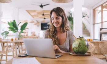 A digital nomad, working remotely on her laptop from a cafe in a tropical location, drinking from a coconut for blog posy how to travel and make money.
