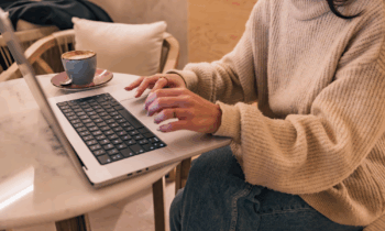 A woman in a bulky sweater working on her laptop in a coffee shop looking for work from home no fee jobs