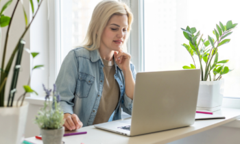 A woman surrounded by plants reading an article on her laptop titled odd ways to make money