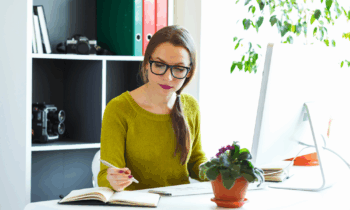 A woman wearing glasses is sitting at her desk looking at her notebook and list of non-phone work from home jobs