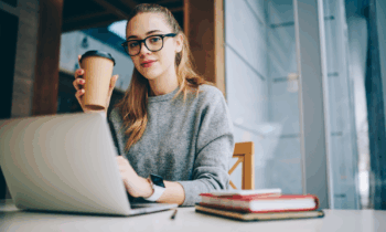 A young woman wearing glasses and drinking coffee looking for work at home jobs with employee benefits on her laptop